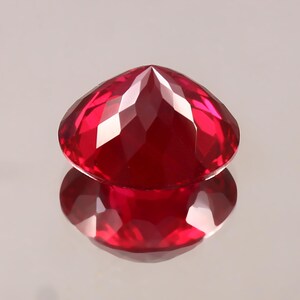 May include: A faceted, heart-shaped ruby gemstone. The gem is a deep, saturated red with multiple facets that reflect light, creating bright highlights. The stone is displayed on a reflective surface, showing its mirrored image.