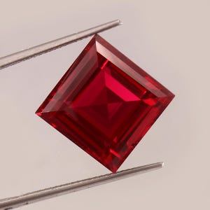 以下が含まれることがあります： ピンセットで保持された、正方形にカットされ、ファセット加工されたルビーの宝石。 宝石は、鮮やかで飽和した赤色で、シャープでクリーンなエッジを持っています。 ファセットは光を反射し、鮮やかな外観を作り出しています。
