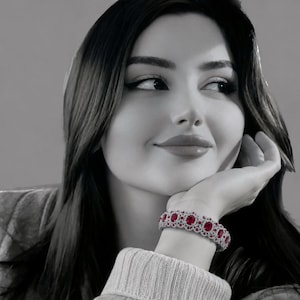 May include: A silver-toned bracelet featuring a series of oval-cut red gemstones surrounded by smaller clear stones. The bracelet is worn on a wrist, with the red and clear stones standing out against the black and white tones of the image.