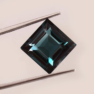 May include: A square-cut, teal-colored gemstone held by tweezers. The gem has a deep, rich color with facets that reflect light. The gemstone is a square shape, and the tweezers are silver.