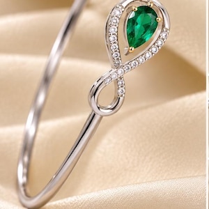 May include: A silver-toned bangle bracelet featuring an infinity symbol centerpiece. The infinity loop is adorned with small clear stones and holds a single pear-shaped green gemstone set in a gold-toned prong setting at the top.