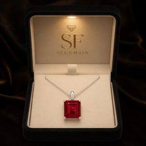 Può includere: Una collana color argento con un ciondolo rettangolare in pietra preziosa rosso intenso. Il gioiello è presentato in una scatola nera foderata in velluto con interno beige, con il testo dorato SF e SFGEMSIN stampato sul coperchio interno.