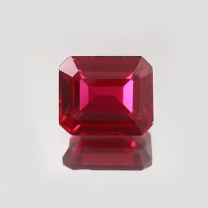 Peut inclure: Une pierre précieuse rubis taillée en forme rectangulaire. La gemme est d'un rouge profond et vibrant, avec une surface réfléchissante, mettant en valeur les facettes internes et une finition polie. Le rubis est posé sur un fond neutre, avec son reflet visible en dessous.