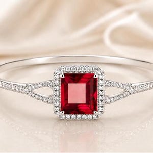 May include: A silver-toned hinged bangle bracelet featuring a central rectangular red gemstone surrounded by a halo of small clear stones. The band splits into two arms on each side of the centre setting, with both arms encrusted with clear stones.