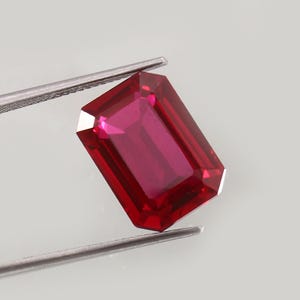 May include: A vibrant, deep red gemstone with an emerald-cut shape, featuring sharp, clean facets that reflect light. The rectangular stone is held securely by a pair of thin, silver-toned metal tweezers, showcasing its clarity and color.