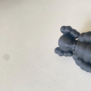 May include: A matte black vinyl collectible figure featuring a rounded head with bone-like ears, X-shaped eyes, and a rounded nose. The figure has a segmented, rounded body structure with visible joints at the shoulders and arms, rendered in a solid dark color.