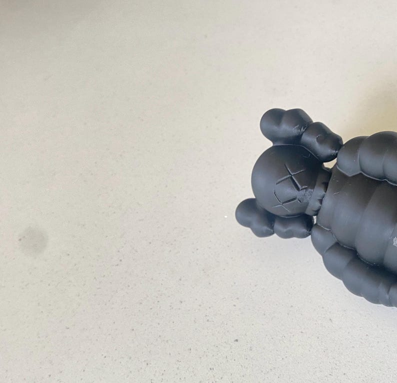 May include: A matte black vinyl collectible figure featuring a rounded head with bone-like ears, X-shaped eyes, and a rounded nose. The figure has a segmented, rounded body structure with visible joints at the shoulders and arms, rendered in a solid dark color.