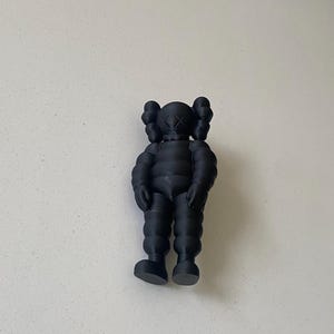May include: A matte black KAWS collectible figure featuring a rounded, segmented body, prominent ears, and signature X-shaped eyes. The character stands in a neutral pose with arms hanging down at its sides, showcasing a smooth, uniform finish.