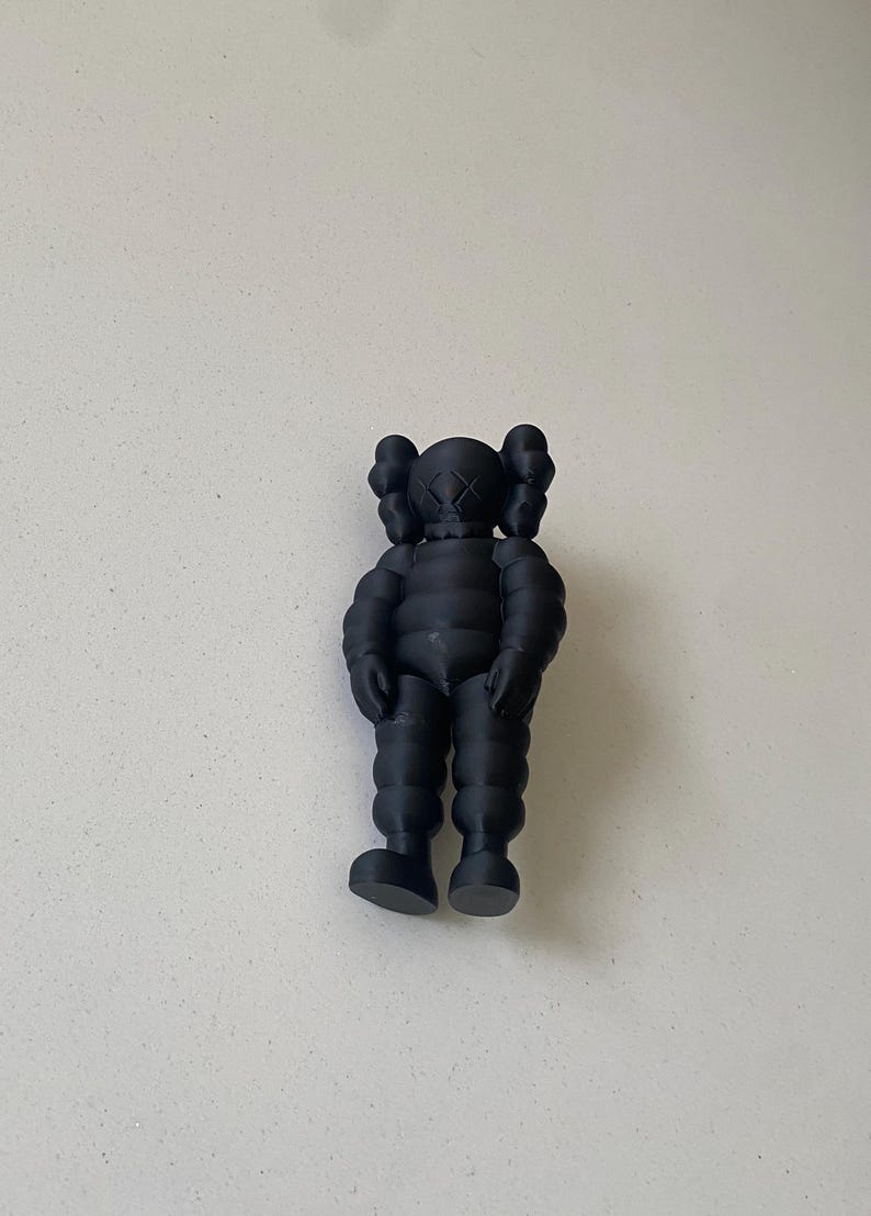 May include: A matte black KAWS collectible figure featuring a rounded, segmented body, prominent ears, and signature X-shaped eyes. The character stands in a neutral pose with arms hanging down at its sides, showcasing a smooth, uniform finish.