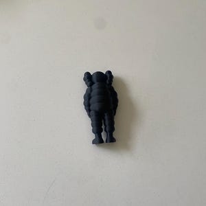 May include: A matte black figurine featuring a stylized character with rounded limbs, a segmented torso, and distinct X-shaped markings on the ears. The figure stands in an upright position with arms resting at its sides, showcasing a smooth, uniform finish.