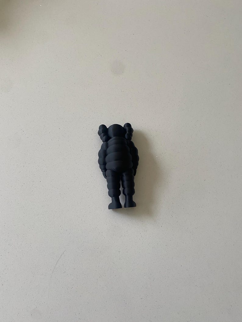 May include: A matte black figurine featuring a stylized character with rounded limbs, a segmented torso, and distinct X-shaped markings on the ears. The figure stands in an upright position with arms resting at its sides, showcasing a smooth, uniform finish.