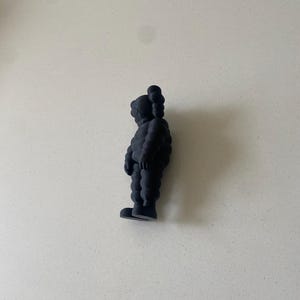 May include: A matte black collectible figure in a standing position, featuring a rounded, segmented body, prominent ears, and an "X" symbol on the side of the head. The figure is shown from a side profile view, displaying its smooth, uniform texture.