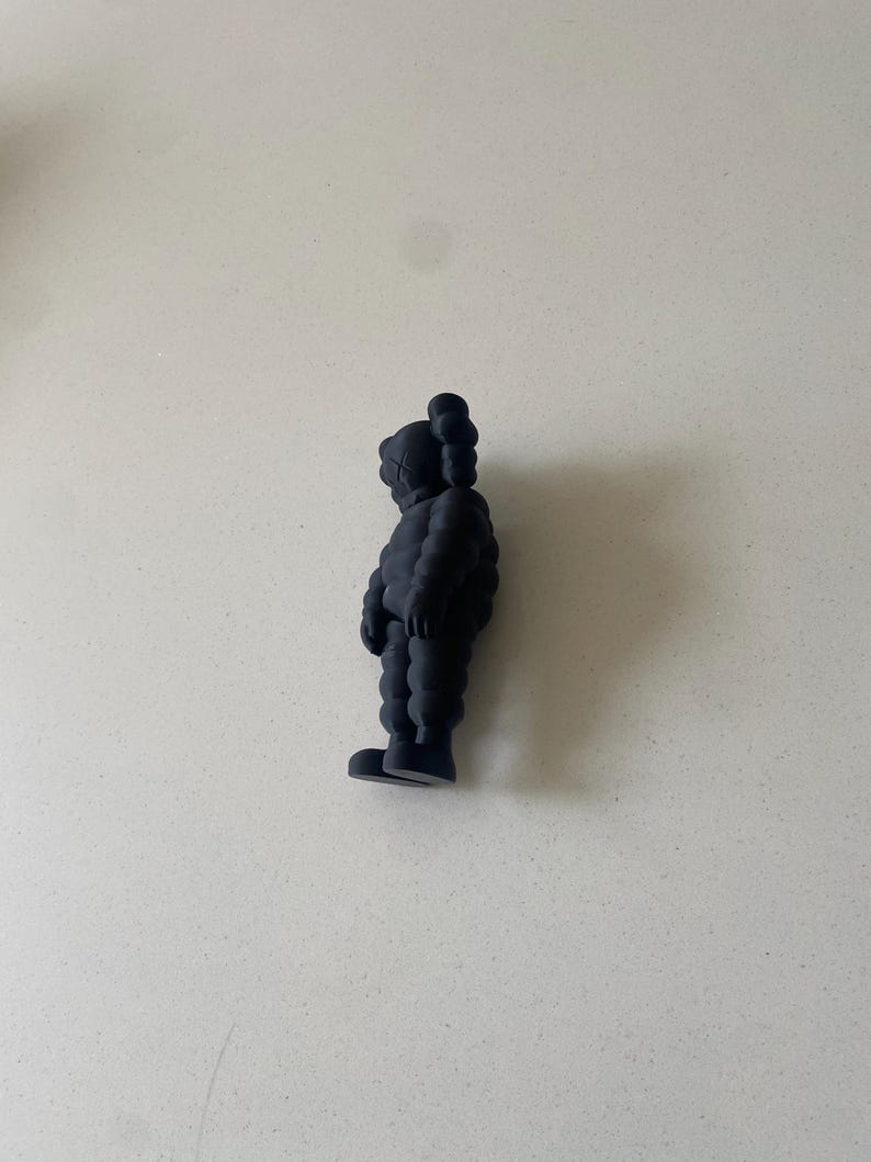 May include: A matte black collectible figure in a standing position, featuring a rounded, segmented body, prominent ears, and an "X" symbol on the side of the head. The figure is shown from a side profile view, displaying its smooth, uniform texture.