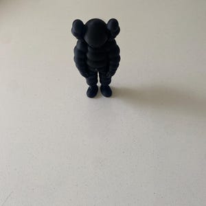 May include: A matte black collectible figure featuring a rounded, segmented body, short limbs, and a head with two prominent, rounded ear-like protrusions. The figure stands in a downward-facing, slumped posture with arms hanging at its sides.