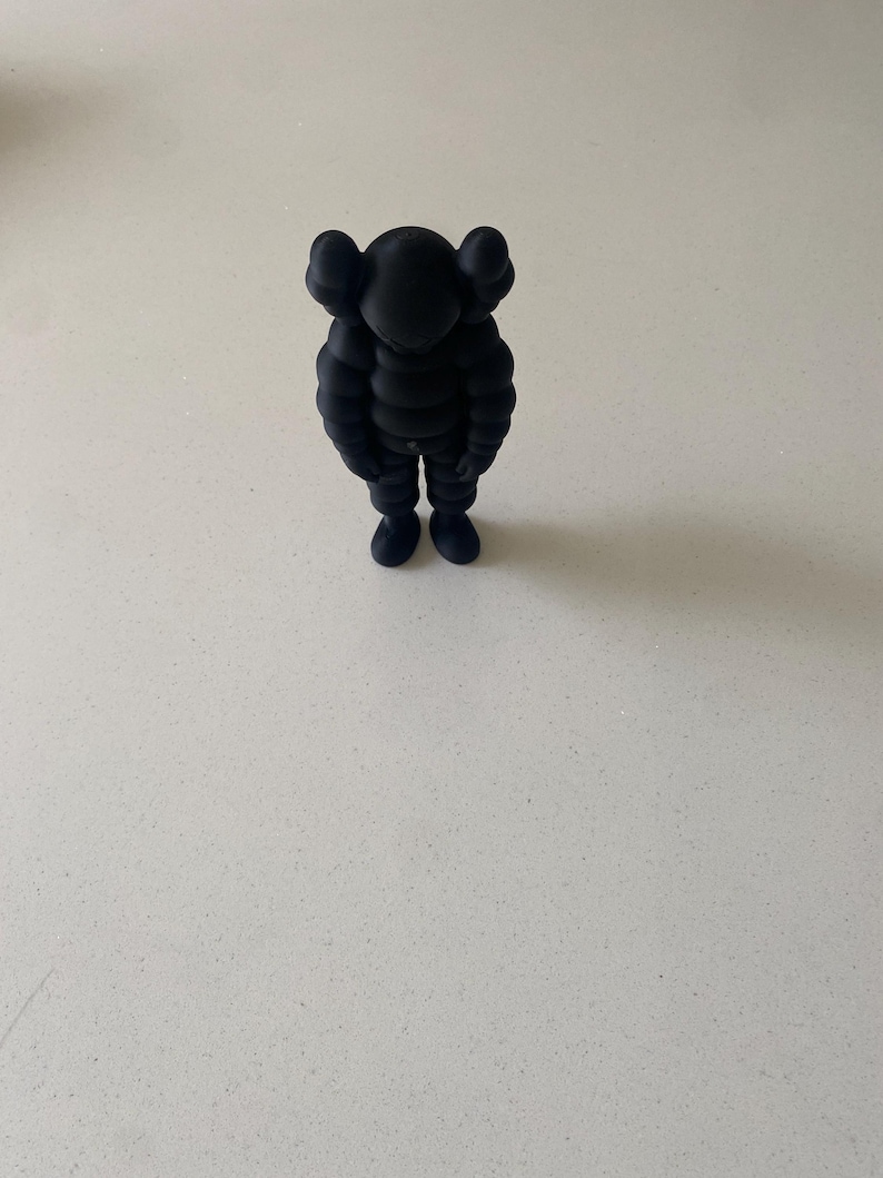 May include: A matte black collectible figure featuring a rounded, segmented body, short limbs, and a head with two prominent, rounded ear-like protrusions. The figure stands in a downward-facing, slumped posture with arms hanging at its sides.