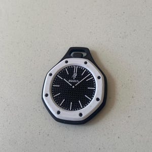 以下が含まれることがあります： 「AP Swatch」の文字が入った白黒の時計チャーム。 時計の文字盤は、白い時間マーカーと針を備えたテクスチャードデザインです。 時計は白と黒のフレームにセットされています。