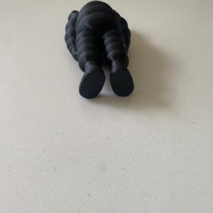 May include: A matte black figurine featuring a segmented, rounded body with a smooth texture. The figure is depicted from a rear-facing perspective, showing two distinct feet and a curved, layered torso design that resembles a stylized character.