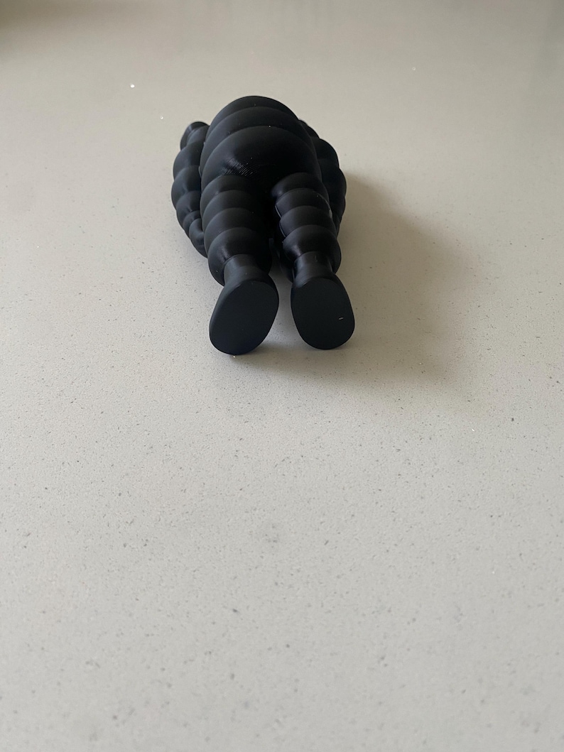 May include: A matte black figurine featuring a segmented, rounded body with a smooth texture. The figure is depicted from a rear-facing perspective, showing two distinct feet and a curved, layered torso design that resembles a stylized character.