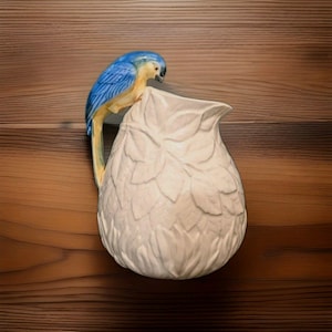 May include: A cream-colored ceramic pitcher featuring a textured surface with embossed leaf patterns. The handle is shaped like a blue and yellow parrot perched on the rim, with its tail feathers extending down the side of the vessel.