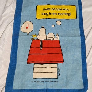 May include: A rectangular blue and light blue tea towel featuring a cartoon of Snoopy lying on a red doghouse roof with Woodstock. A yellow speech bubble contains the text I hate people who sing in the morning! with a musical note icon nearby.