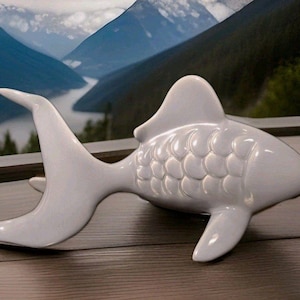 May include: A white ceramic fish figurine with a smooth, glossy finish. The sculpture features a prominent dorsal fin, a wide tail, and molded scale details along the body. The fish is positioned horizontally, showcasing its curved, fluid shape.