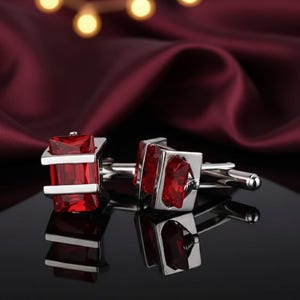 May include: A pair of silver cufflinks with a rectangular, ruby-red gemstone center. The cufflinks have a square frame and a T-bar closure. The background is a deep burgundy fabric with soft lighting.