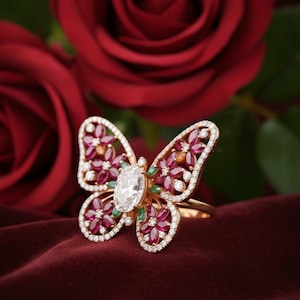 May include: A gold butterfly ring with a large oval-cut diamond center stone. The butterfly wings are adorned with red and green gemstones, and outlined with small white diamonds. The ring is set against a backdrop of red roses.