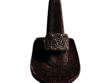 Puede incluir: Un anillo plateado con un diseño texturizado en forma de ola, engastado con pequeñas piedras oscuras. El anillo se exhibe en un soporte de anillo de terciopelo marrón oscuro sobre un fondo blanco. Se destacan los intrincados detalles del anillo.