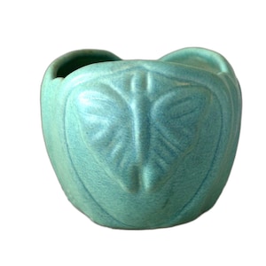 May include: A small, round, turquoise ceramic planter featuring a raised, embossed butterfly design on the front. The vessel has a textured, matte finish and a wide, open top with a scalloped rim, showcasing a consistent light blue-green color.