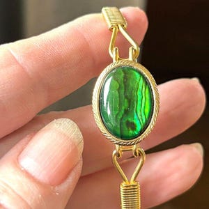 May include: A gold-toned bracelet featuring a central oval-shaped green iridescent stone set in a rope-textured bezel. The bracelet band is constructed from gold-colored wire, with decorative wire-wrapped accents at the connection points.
