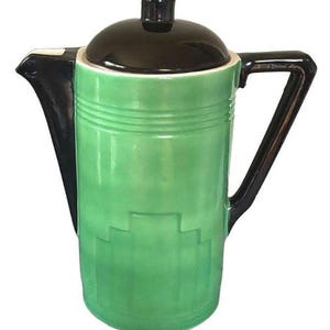 May include: A ceramic coffee pot featuring a bright green cylindrical body with a stepped geometric relief pattern near the base. It includes a glossy black lid, a black angular handle, and a black spout, creating a distinct contrast in color.