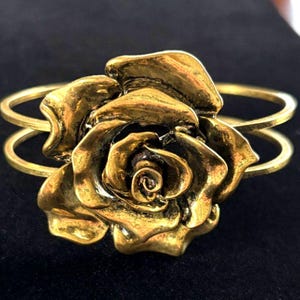 May include: A gold-toned cuff bracelet featuring a detailed rose design. The rose is the focal point, with textured petals and a central spiral. The bracelet has a double-band design for a secure fit and is set against a dark background.
