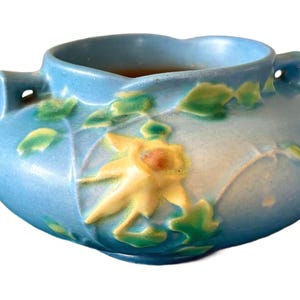May include: A light blue ceramic bowl with two small handles on the sides. The exterior features a raised relief design of a yellow flower with green leaves. The interior of the bowl is a dark brown color, and the rim has a scalloped edge.