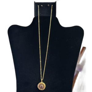 May include: A gold-colored necklace with a round pendant featuring a pattern of small, dark red gemstones. The pendant hangs from a delicate chain. The necklace is displayed on a black mannequin.