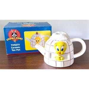 Puede incluir: Tetera de cerámica blanca de Tweety Bird con una figura amarilla de Tweety Bird en la parte delantera. La tetera tiene un asa y un pico blancos. La caja detrás de la tetera tiene el logotipo de Looney Tunes y las palabras "Tweety Ceramic Tea Pot."