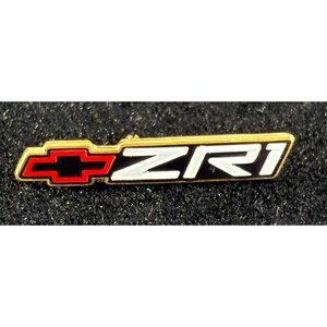 Puede incluir: Un pin dorado con el logotipo de Chevrolet y las letras "ZR1" en blanco. El logotipo de Chevrolet es rojo y negro. El pin es rectangular y está sobre un fondo negro texturizado.
