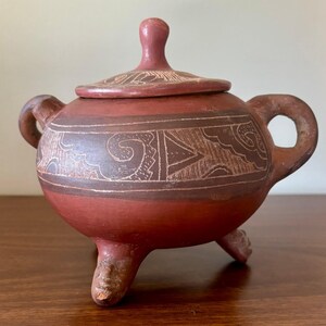 Puede incluir: Un recipiente de cerámica color terracota con tapa, que cuenta con dos asas laterales y tres patas. El cuerpo y la tapa presentan patrones geométricos grabados. La pata frontal está esculpida con la forma de un rostro pequeño, acabado mate.