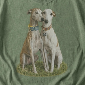 Puede incluir: Una camiseta de manga corta color verde oliva con un estampado gráfico de dos perros sentados en la hierba. El perro de la izquierda es marrón y blanco con un collar azul, y el de la derecha es blanco y beige con un collar marrón. Cuello redondo.
