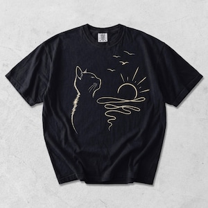May include: A black short-sleeved t-shirt featuring a cream-coloured line art graphic on the front. The design depicts the profile of a cat looking towards a rising sun over wavy water, with three small birds flying in the sky above the sun.
