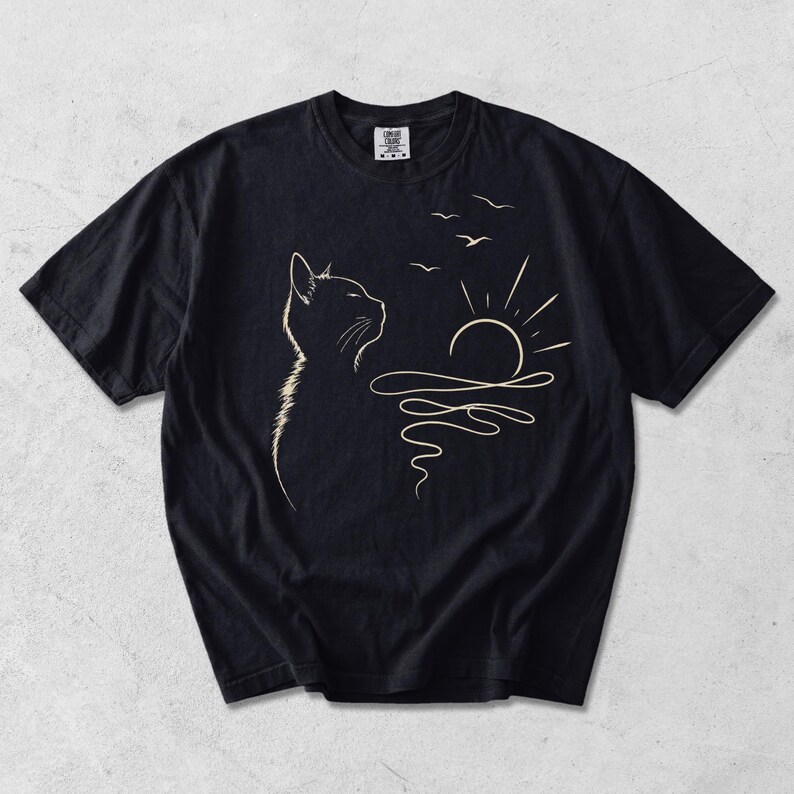 May include: A black short-sleeved t-shirt featuring a cream-coloured line art graphic on the front. The design depicts the profile of a cat looking towards a rising sun over wavy water, with three small birds flying in the sky above the sun.
