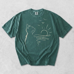 May include: A dark green short-sleeved t-shirt featuring a light-coloured line art graphic of a cat profile looking towards a sun rising over wavy water with three birds flying above. The shirt has a crew neckline and a visible brand label at the neck.