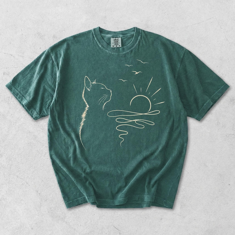 May include: A dark green short-sleeved t-shirt featuring a light-coloured line art graphic of a cat profile looking towards a sun rising over wavy water with three birds flying above. The shirt has a crew neckline and a visible brand label at the neck.