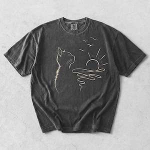 May include: A charcoal grey short-sleeve t-shirt featuring a light-coloured line art graphic of a cat looking towards a rising sun over water with three birds flying above. The shirt has a crew neckline and a visible brand tag at the inner collar.