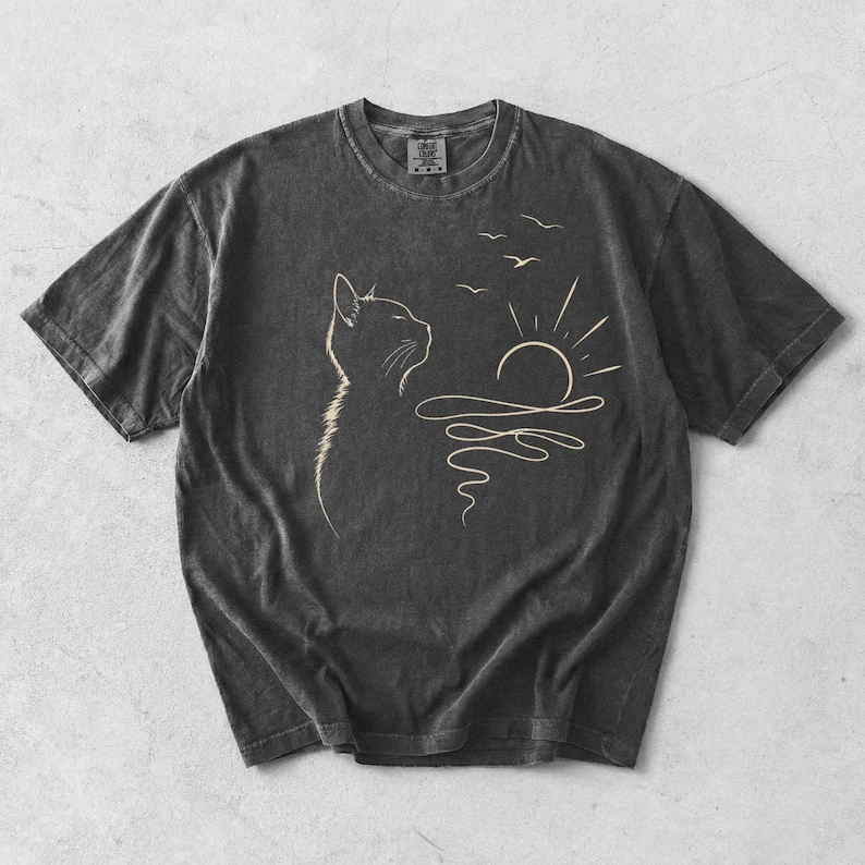 May include: A charcoal grey short-sleeve t-shirt featuring a light-coloured line art graphic of a cat looking towards a rising sun over water with three birds flying above. The shirt has a crew neckline and a visible brand tag at the inner collar.