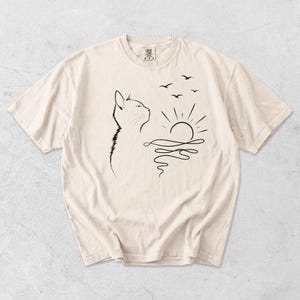 May include: A cream-coloured short-sleeve t-shirt featuring a black line art graphic on the front. The design depicts the profile of a cat looking towards a rising sun over water, with three small birds flying in the sky above the sun.