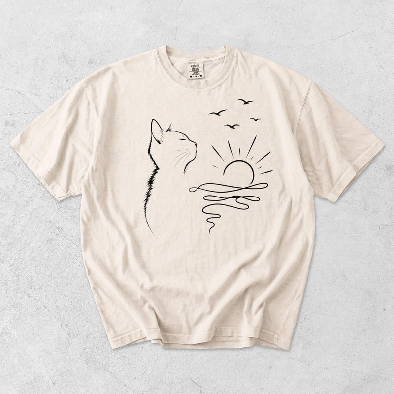 May include: A cream-coloured short-sleeve t-shirt featuring a black line art graphic on the front. The design depicts the profile of a cat looking towards a rising sun over water, with three small birds flying in the sky above the sun.