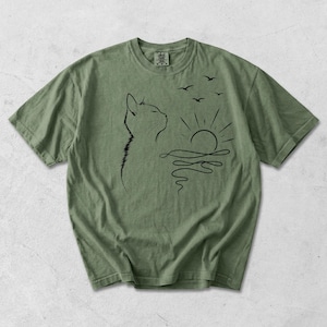 May include: An olive green short-sleeve t-shirt featuring a black line art graphic of a cat in profile looking towards a stylised sun rising over wavy water with three small birds flying above. The shirt has a crew neckline and a visible brand tag.