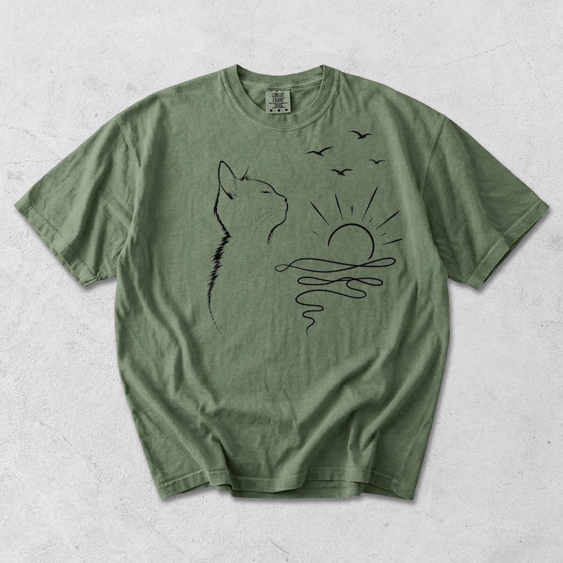May include: An olive green short-sleeve t-shirt featuring a black line art graphic of a cat in profile looking towards a stylised sun rising over wavy water with three small birds flying above. The shirt has a crew neckline and a visible brand tag.
