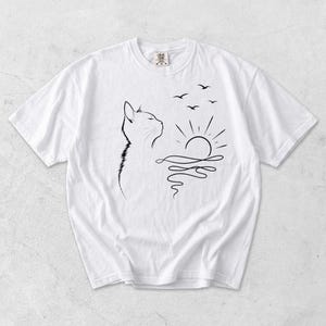 May include: A white short-sleeved t-shirt featuring a black line art graphic on the front. The design depicts the profile of a cat looking towards a rising sun over wavy water, with three small birds flying in the sky above the sun.
