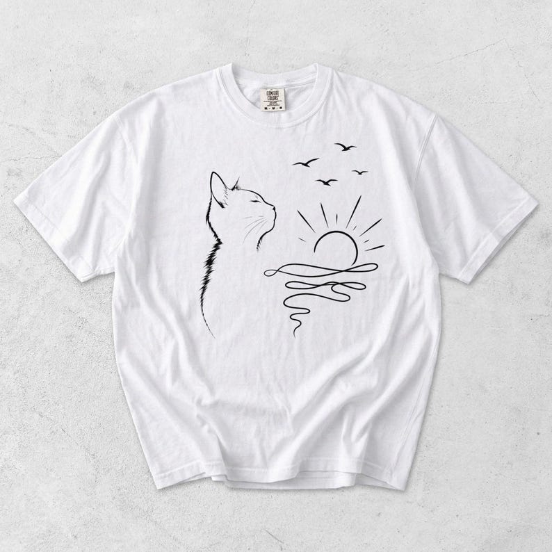 May include: A white short-sleeved t-shirt featuring a black line art graphic on the front. The design depicts the profile of a cat looking towards a rising sun over wavy water, with three small birds flying in the sky above the sun.